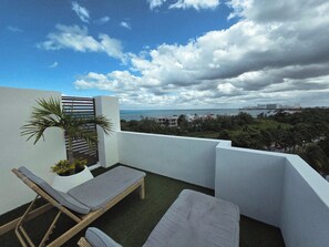 Ein Balkon mit zwei Liegestühlen, einer kleinen Palme und Blick auf das Meer und die Stadtlandschaft.
