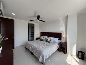 Une chambre à coucher avec un lit, des tables de chevet, un ventilateur de plafond, une porte et une plante en pot.