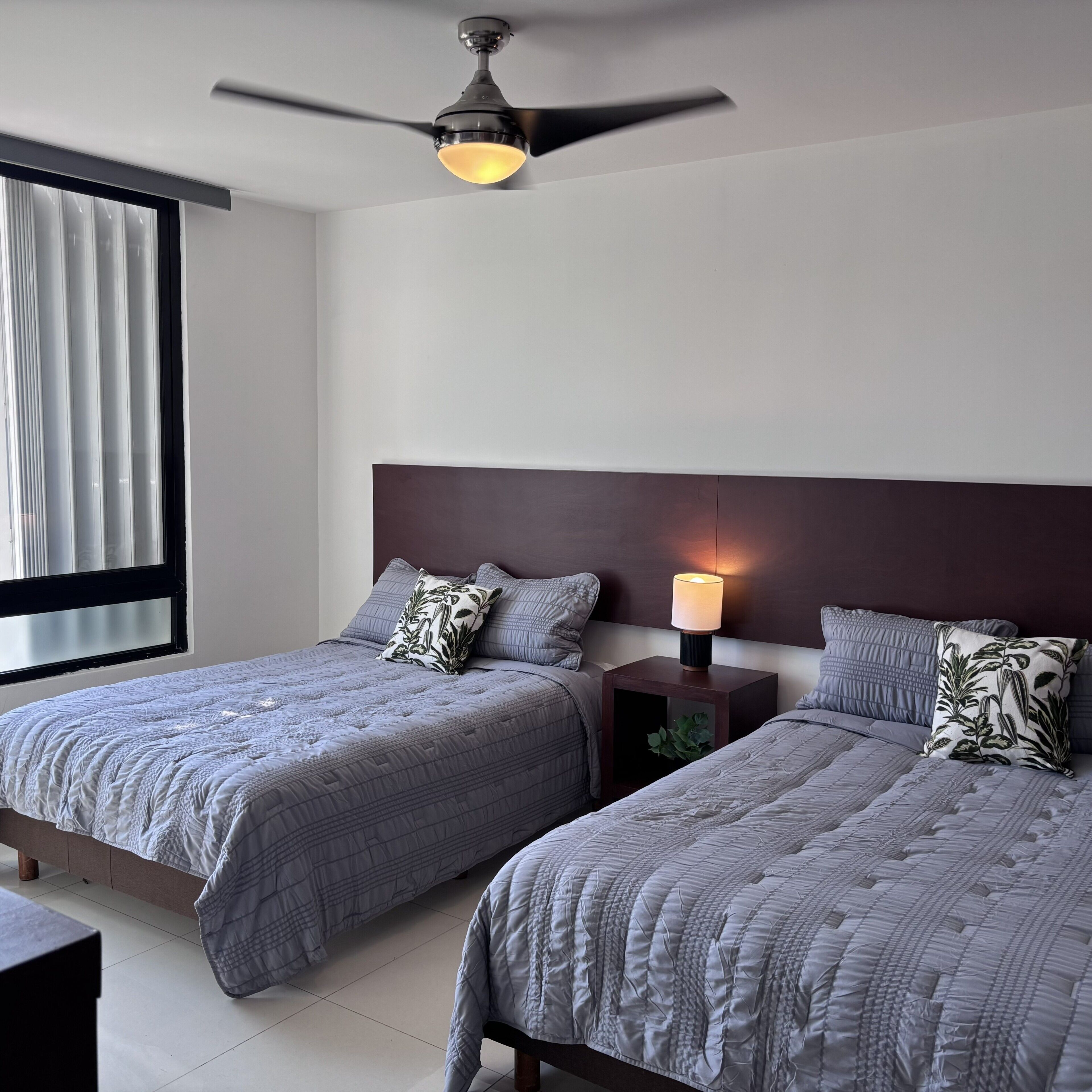 A hotel room with two beds, a ceiling fan, a bedside table with a lamp, and a window with curtains.