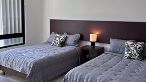 Quarto de hotel com duas camas, um ventilador de teto, uma mesa de cabeceira com abajur e uma janela com cortinas.