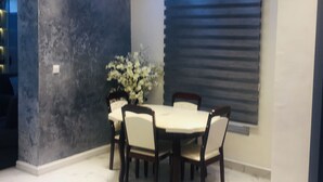 Uma área de refeições com uma mesa redonda, cadeiras, um vaso de plantas e uma janela com persianas.