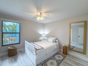 Ein Schlafzimmer mit einem Bett, einem Deckenventilator, einem Fenster mit Ausblick, einem Nachttisch und einem Spiegel.