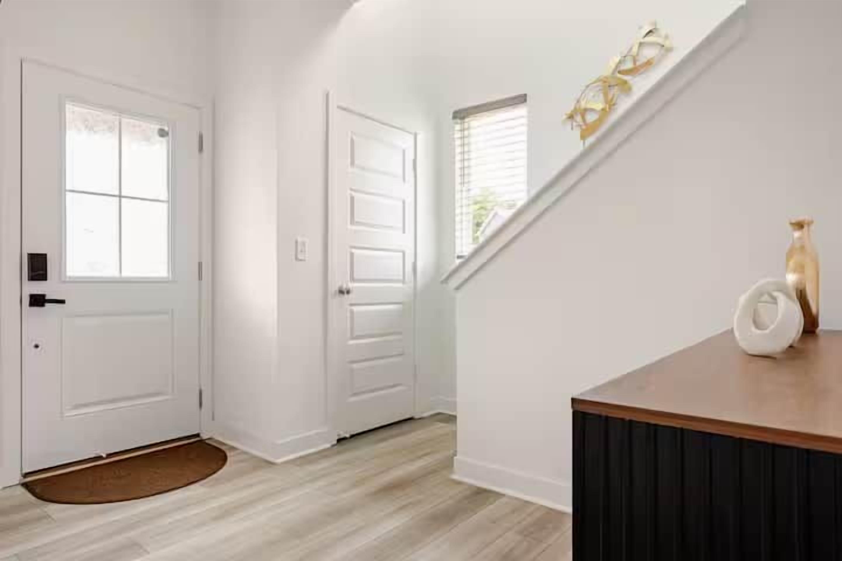 A hallway with a wooden floor, white walls, a door with a window, and a small rug.