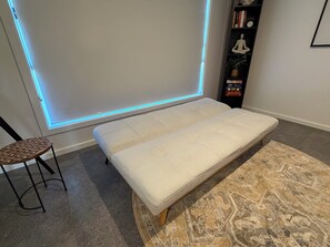 A room with a white cushioned bench, a patterned rug, a small stool, and a bookshelf with books and a clock. - Casa Oliva | Spacious Retreat Near Toranga Zoo (Dubbo)