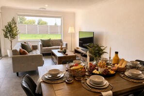 A dining area with a wooden table set for a meal, a sofa, a TV, and a window with a view of a grassy yard. - Casa Oliva | Spacious Retreat Near Toranga Zoo (Dubbo)