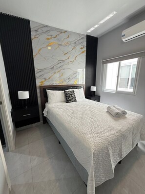 Ein modernes Schlafzimmer mit Wand in Marmoroptik, einem großen Bett mit gesteppter Bettdecke, einem Nachttisch mit Lampe und einem Fenster mit Jalousien.