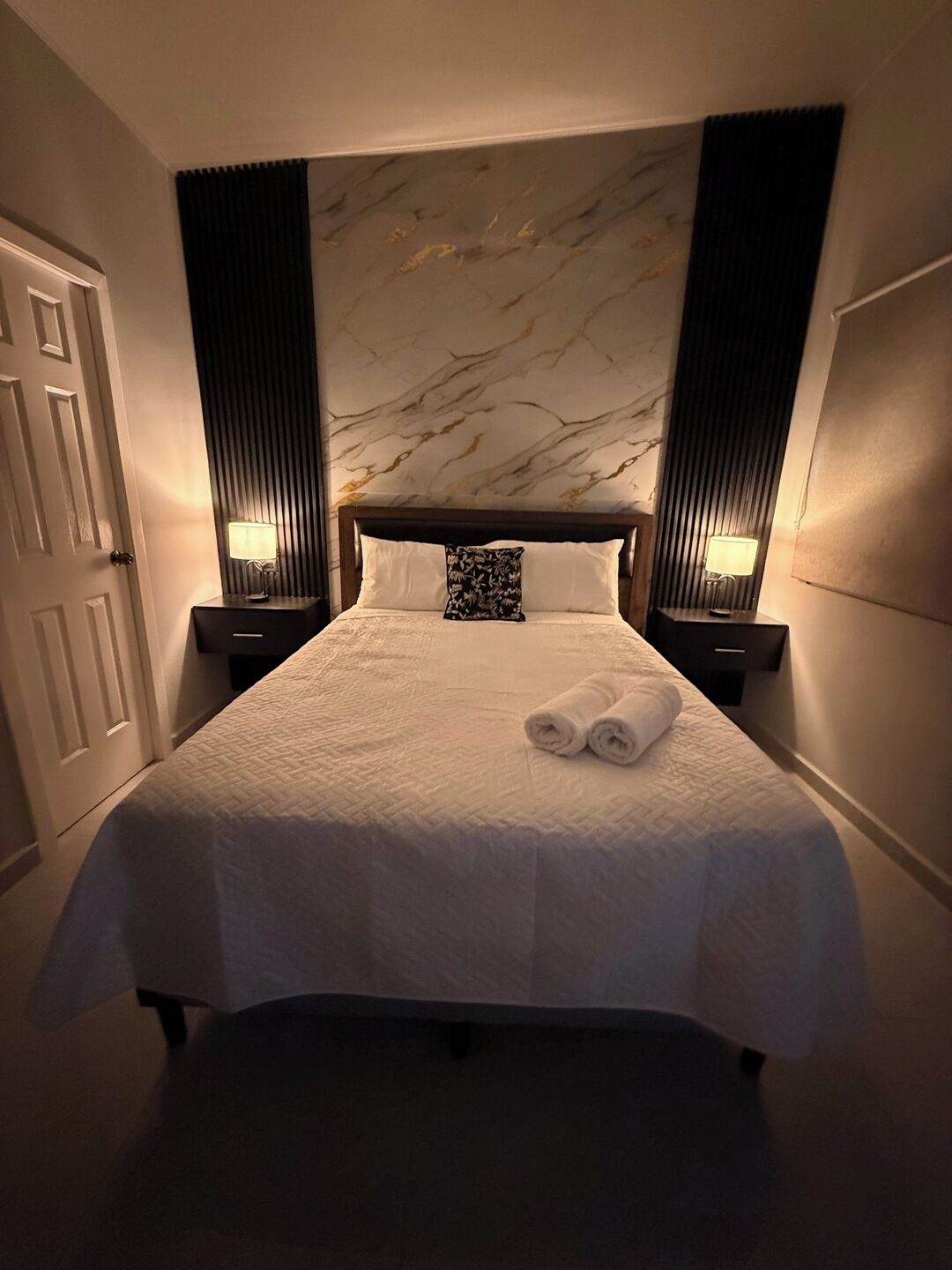 Una habitación de hotel con una cama grande, dos mesitas de noche con lámparas, un cabecero con diseño de mármol y una puerta.