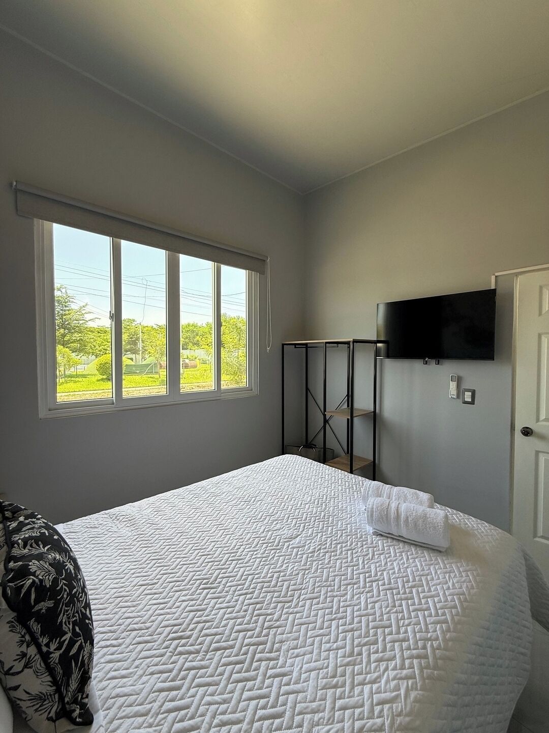 Een slaapkamer met een bed, een aan de muur bevestigde televisie, een raam met uitzicht op groen, en een deur.