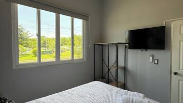 Een slaapkamer met een bed, een aan de muur bevestigde televisie, een raam met uitzicht op groen, en een deur.