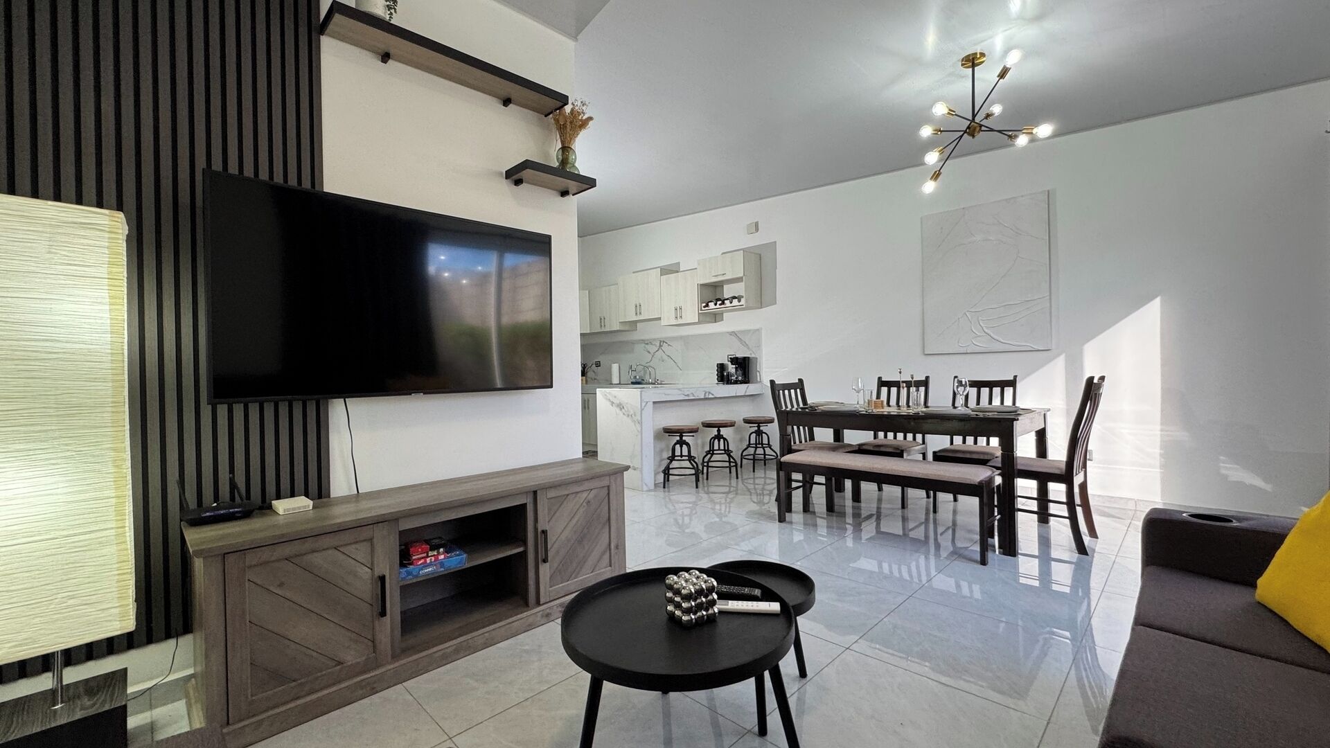 Una sala de estar moderna con televisor de pantalla plana, un comedor con mesa y sillas, y una cocina al fondo.
