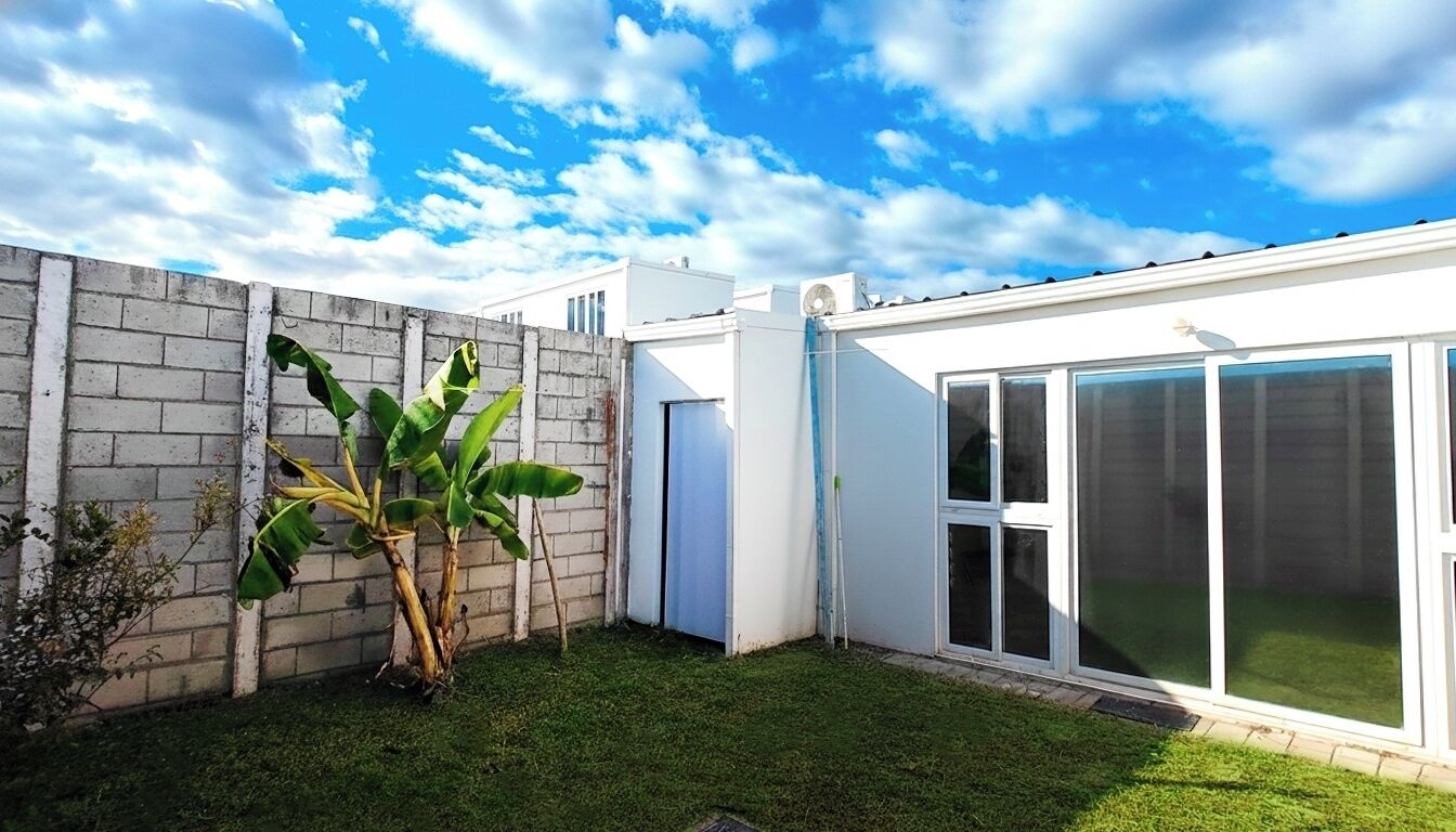 A residential property with a brick wall, a large banana plant, a white building with a sliding glass door, and a clear blue sky.