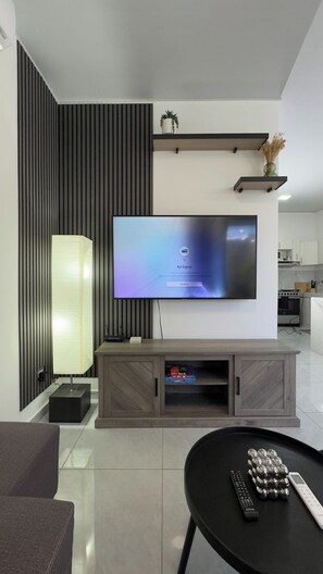 Una sala moderna con un televisor de pantalla plana montado en la pared, un mueble bajo para equipo de sonido de madera, una lámpara de pie y una mesa de centro con controles remotos.