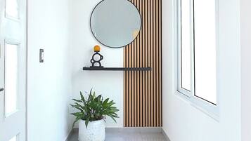 A hallway with a white tiled floor, a potted plant, a mirror on a shelf, and a vertical wooden panel.