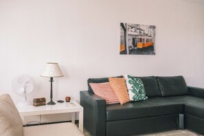 Ein Wohnzimmer mit einem schwarzen Ledersofa, einem weißen Beistelltisch mit Lampe, einem Ventilator und einem kleinen Dekorationsgegenstand.