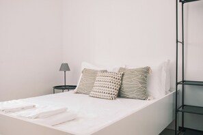 Un lit simple avec une literie blanche, deux coussins à motifs, une petite table ronde avec une lampe et une étagère en métal.