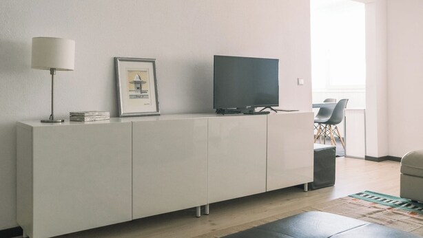 En moderne stue med et hvidt skab, et fladskærms-tv, et indrammet billede, en gulvlampe og et spiseområde med stole.