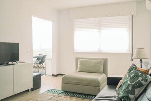 Un salon moderne comprenant un canapé, un téléviseur, un coin repas avec des chaises et une fenêtre avec des stores.