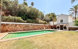 A swimming pool with a stone wall, a grassy lawn, and a white building with a balcony.