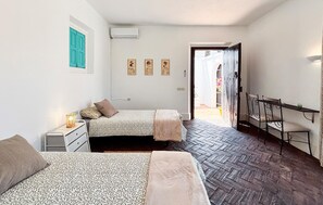A room with two beds, a small table with a lamp, a wardrobe, and a window with a blue shutter.
