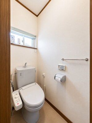 Casa de banho pequena e limpa, com sanita, secador de mãos, dispensador de papel e janela com barra de toalhas.
