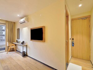 A hotel room with a wooden door, a flat-screen TV mounted on the wall, a small desk with a chair, and an air conditioning unit. - Palette Hotel Millennium City Gurugram (Gurugram)