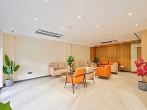Ein modernes Wohnzimmer mit einem beigen Sofa, orangefarbenen Sesseln und einem Couchtisch. Im Hintergrund sind Kübelpflanzen und eine Holzkommode zu sehen.