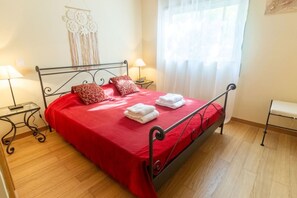 Ein Bett mit roter Tagesdecke, zwei Kissen und zwei gefaltete Handtücher. Ein Nachttisch mit Lampe, einer Wanddekoration und einem Fenster mit durchsichtigen Vorhängen.