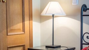 Quarto de hotel com porta de madeira, mesa de cabeceira com abajur, cama com manta vermelha e travesseiro vermelho.