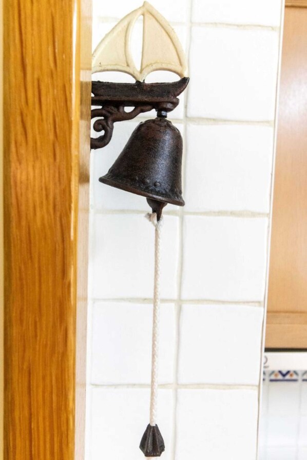Una campana de metal negro con una cuerda colgando, fijada a una pared con fondo de azulejos blancos.