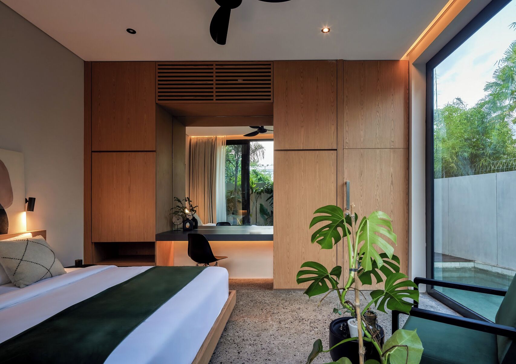 Un dormitorio moderno con cama, mesita de noche, lámpara y un amplio ventanal con vistas a la vegetación.