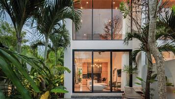 A modern house with a glass facade, a patio with seating, and lush greenery.