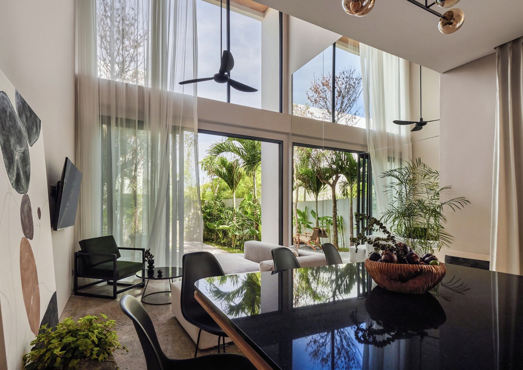 A modern dining area with a glass table, black chairs, and a view of a lush garden through large windows.