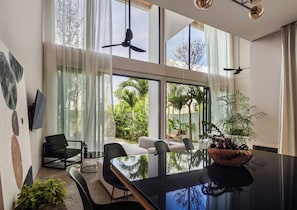 Uma área de refeições moderna com mesa de vidro, cadeiras pretas e vista para um jardim exuberante através de grandes janelas.