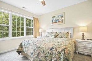 Een slaapkamer met een groot bed, nachtkastje, lamp, raam met gordijnen en een schilderij aan de muur.