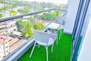 A balcony with a glass railing, two wicker chairs, a small round table, and artificial green grass. - Modern 1BDRM Apartment|Kilimani|Fast WiFi and Cool views of Nairobi (Nairobi)
