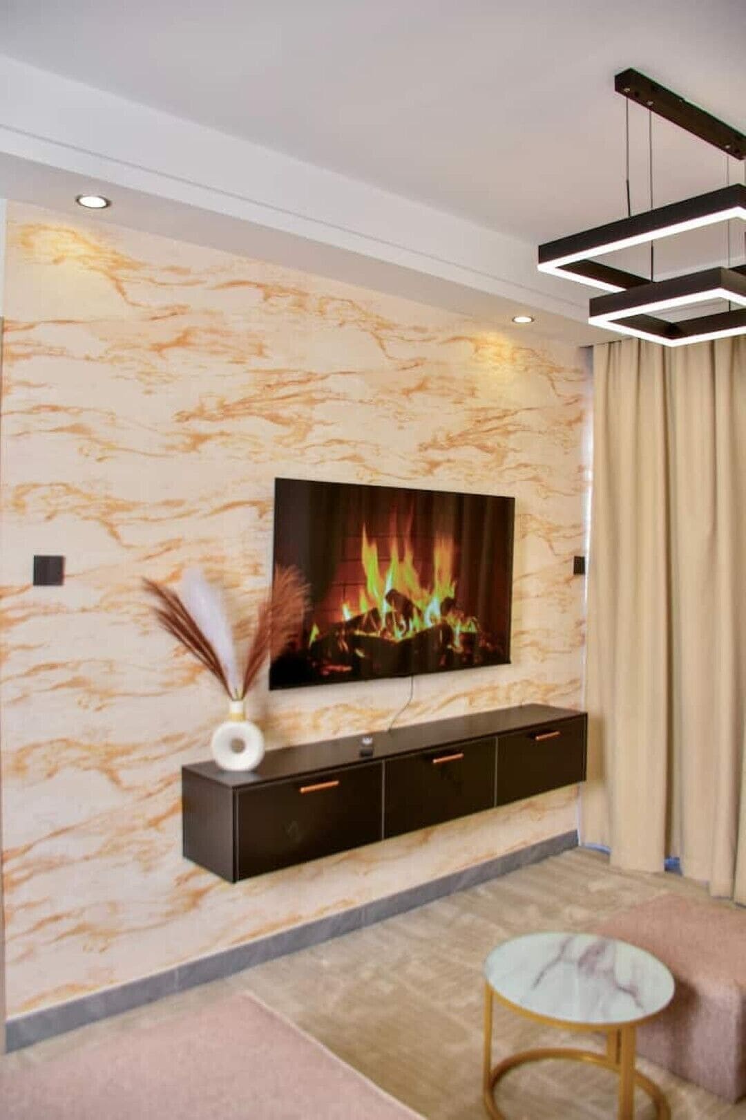 A living room with a flat-screen TV mounted on a marble wall, a dark entertainment unit below, a round coffee table, and a modern pendant light fixture.