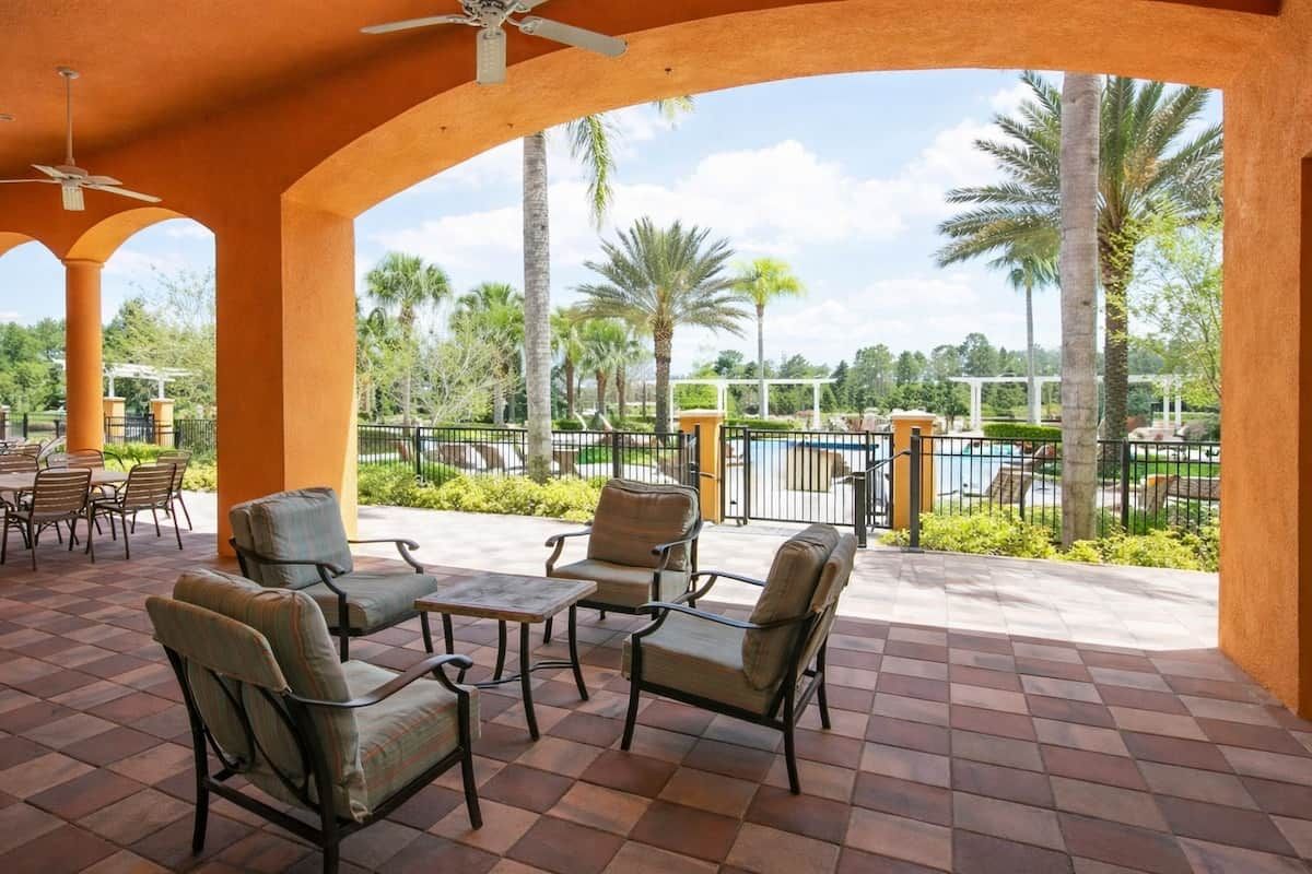 A covered patio area with outdoor seating, a pool, and palm trees in the background.