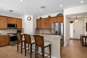 A kitchen with wooden cabinets, stainless steel appliances, a central island with bar stools, and a clock on the wall. - DisneyMagic!Luxury Getaway (Davenport)