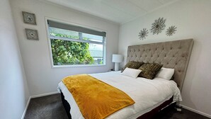 A bedroom with a large bed, a window with a view of greenery, and a decorative wall piece. - Views @ Four Winds (Taupō)