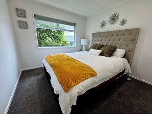 A bedroom with a large bed, a window with a view of greenery, and a decorative wall piece. - Views @ Four Winds (Taupō)