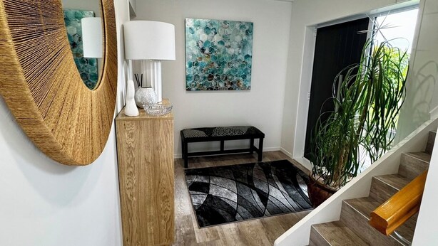A modern interior with a wooden floor, a round mirror, a bench, a painting, and a staircase. - Views @ Four Winds (Taupō)