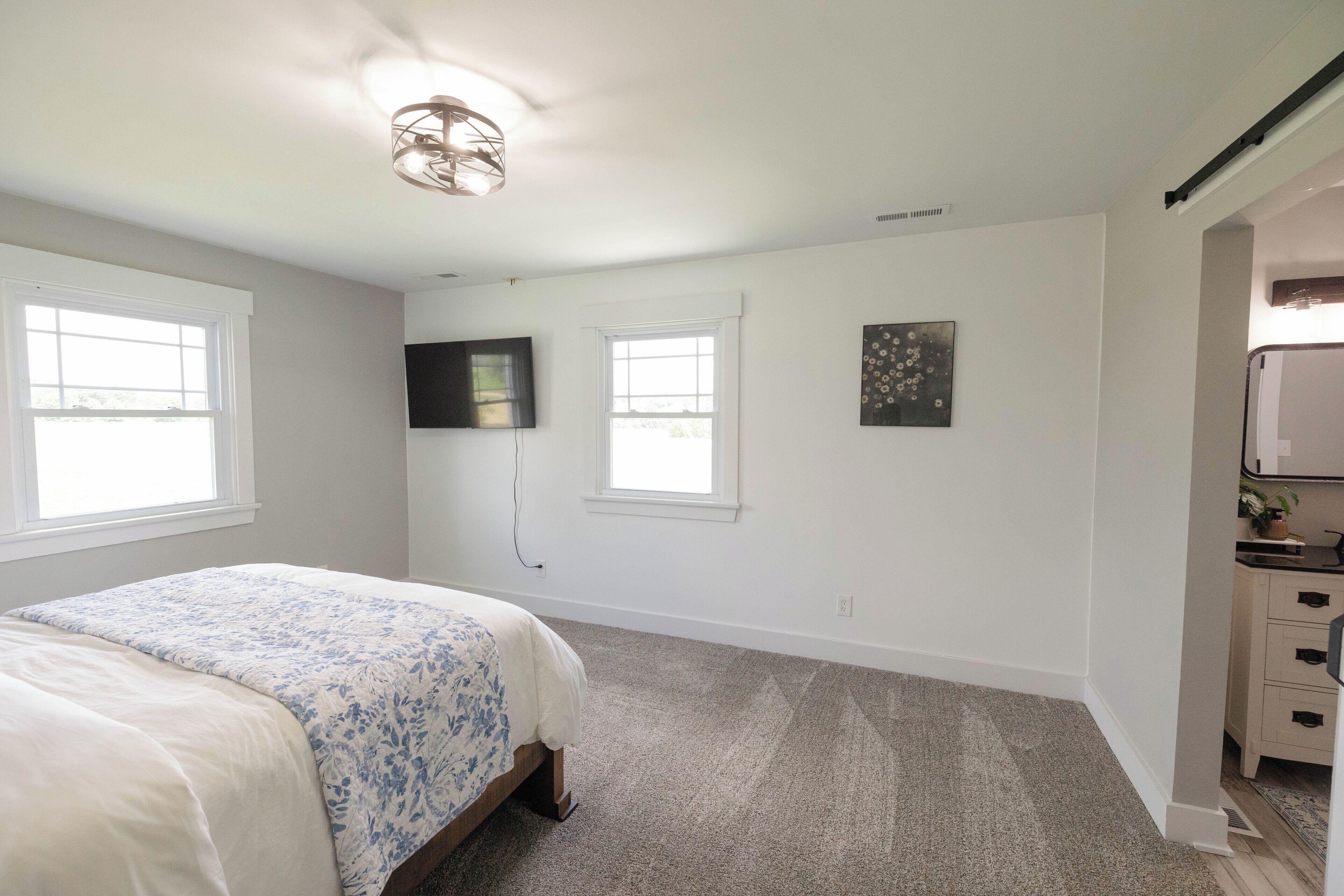 A bedroom with a bed, two windows, a flat-screen TV, and a small artwork on the wall.