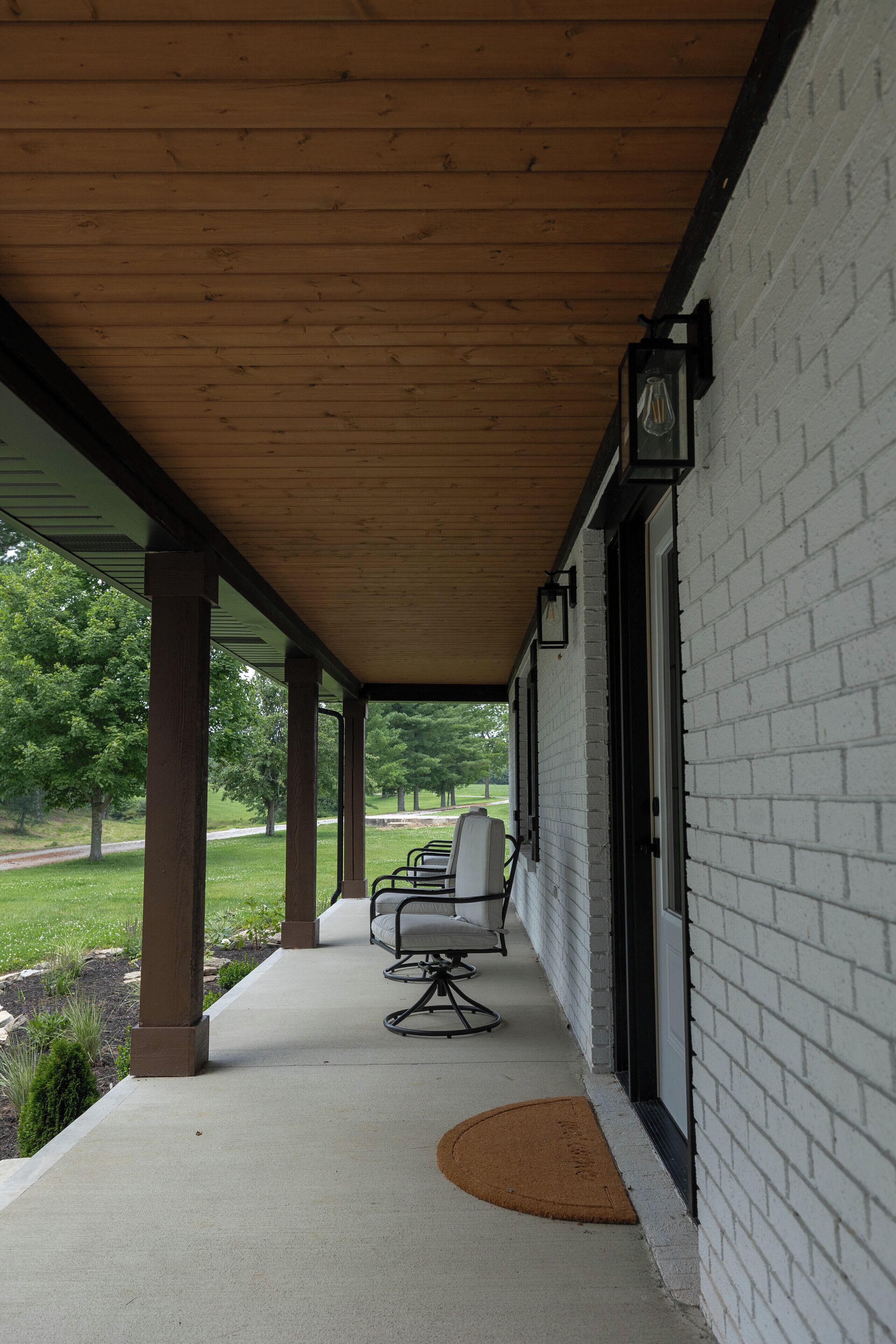 A covered porch with a brick wall, a wooden ceiling, and a single chair.