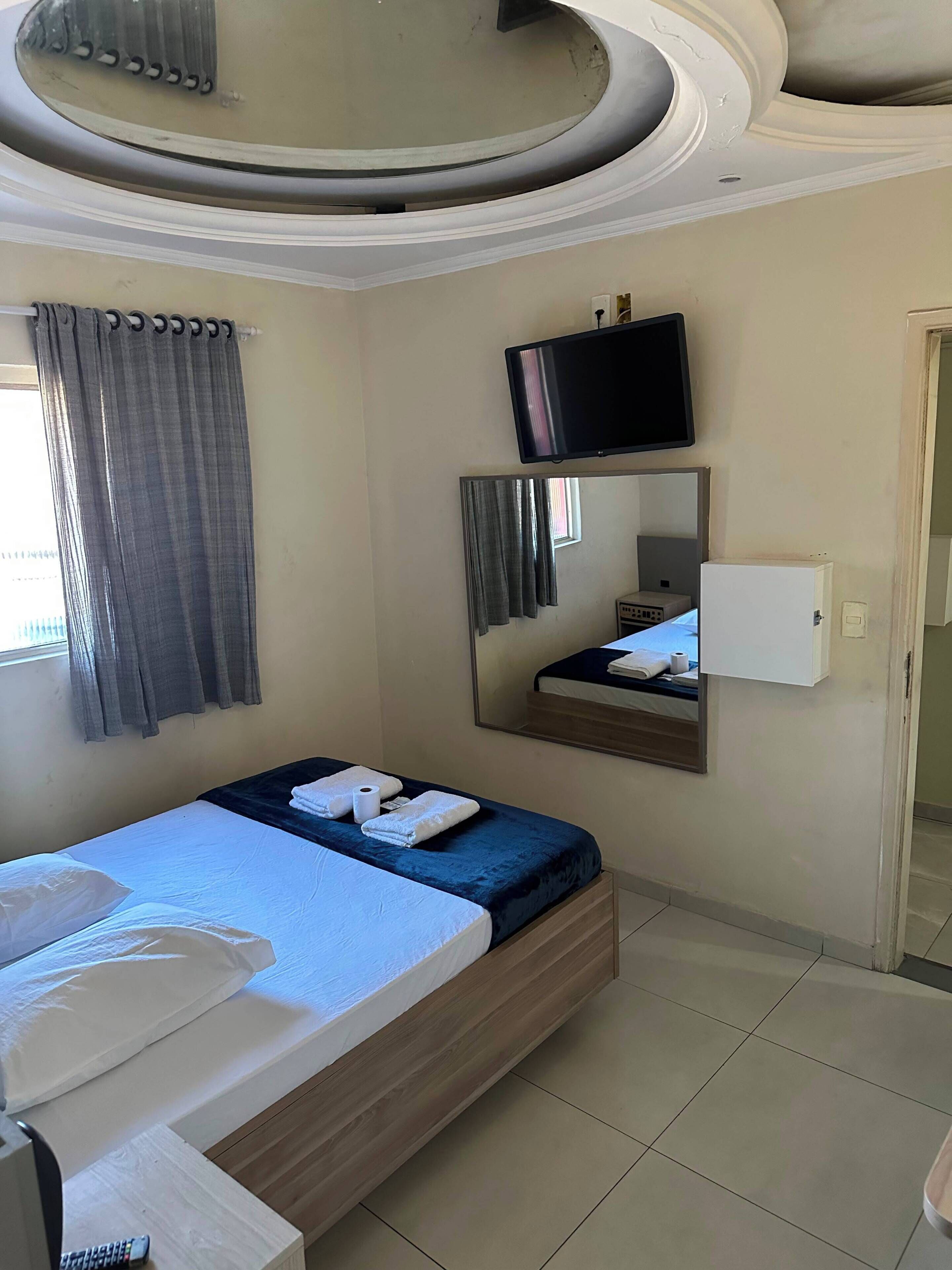 Quarto de hotel com uma cama, televisão, espelho e janela com cortinas.