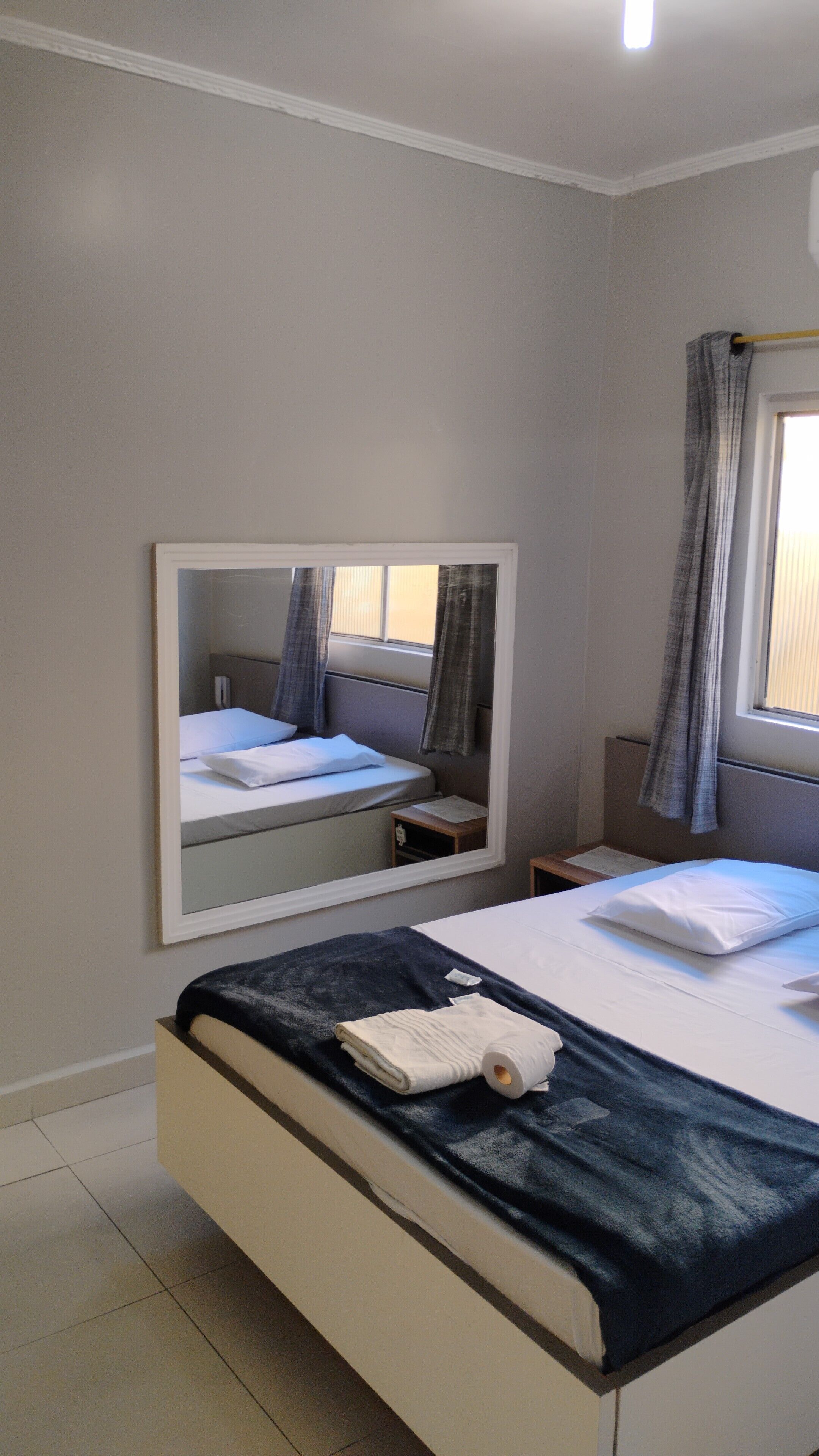 A bedroom with a bed, a bedside table, a mirror, and a window with curtains.