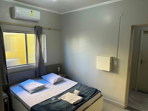 A hotel room with a bed, a window with blinds, an air conditioning unit, and a bedside table with a phone.