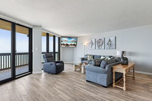A spacious living room with a balcony, a leather recliner, a grey sofa, and a wooden coffee table. - Sandpiper 1005: Oceanfront 3BR condo with pool, hot tub, and beach access in Port Aransas. (Port Aransas)