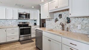 A modern kitchen with white cabinetry, stainless steel appliances, and a herringbone tile backsplash. - Sandpiper 1005: Oceanfront 3BR condo with pool, hot tub, and beach access in Port Aransas. (Port Aransas)