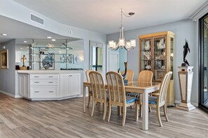 A dining area with a glass-top table, wooden chairs, and a chandelier. - Sandpiper 1005: Oceanfront 3BR condo with pool, hot tub, and beach access in Port Aransas. (Port Aransas)