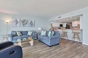 A modern living room with two sofas, a glass coffee table, and a dining area with wooden chairs. - Sandpiper 1005: Oceanfront 3BR condo with pool, hot tub, and beach access in Port Aransas. (Port Aransas)